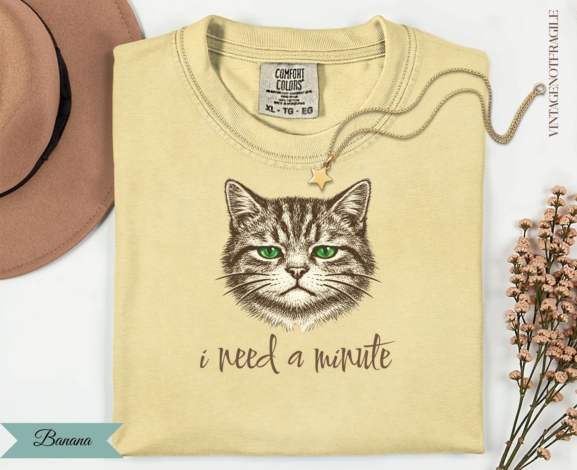 Cream t-shirt with a green-eyed cat illustration and "I need a minute" text.