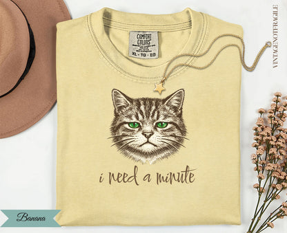 Cream t-shirt with a green-eyed cat illustration and "I need a minute" text.