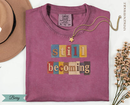 Berry colored t-shirt with "still becoming" text in block letters.