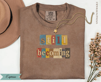 Espresso t-shirt with colorful "still becoming" text graphic.