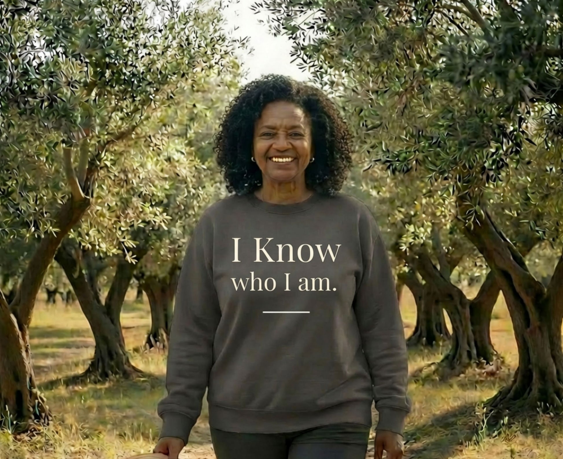 Smiling woman in olive grove wearing sweatshirt with "I Know Who I Am."