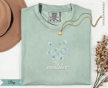 Pale green t-shirt with blue flowers and "muhler" text, with gold necklace.