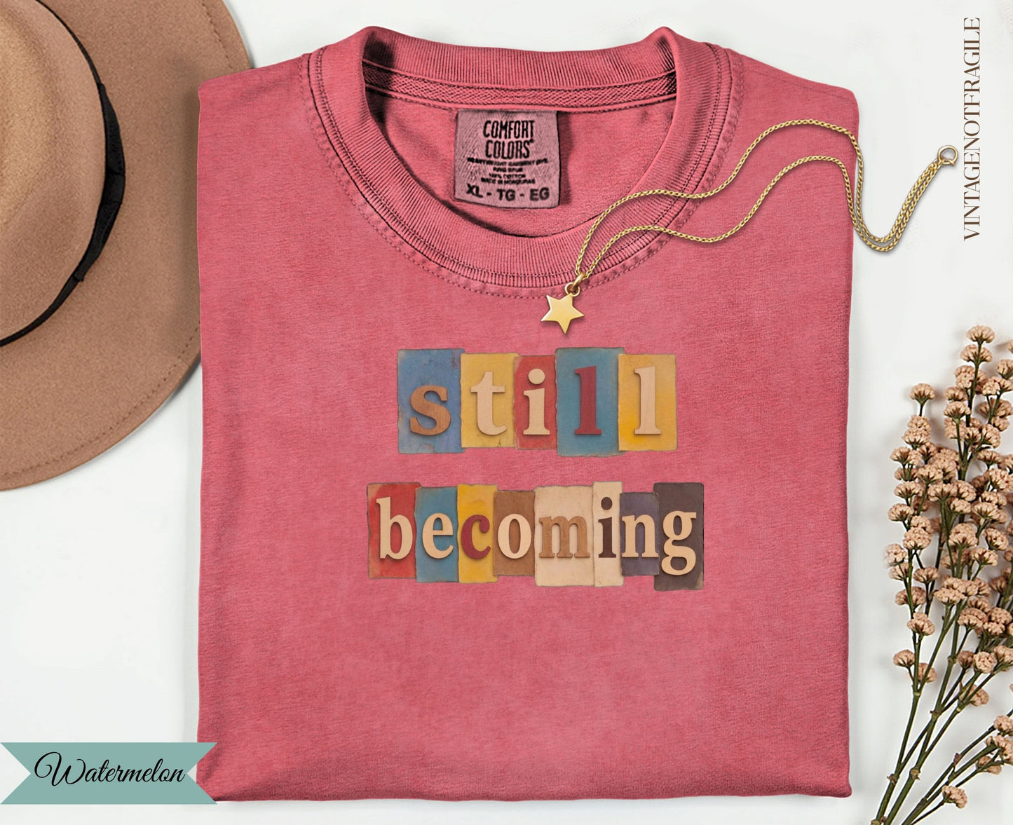 Still Becoming text graphic on a pink t-shirt with gold necklace and hat.