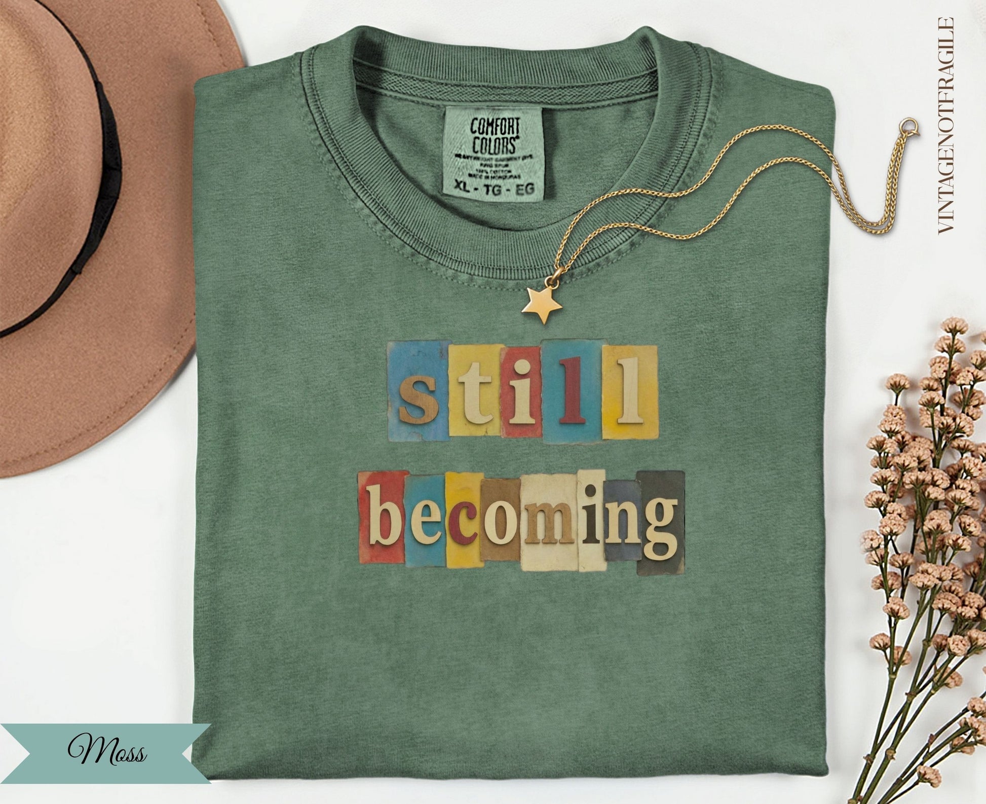 Green t-shirt with "still becoming" text in colorful block letters.