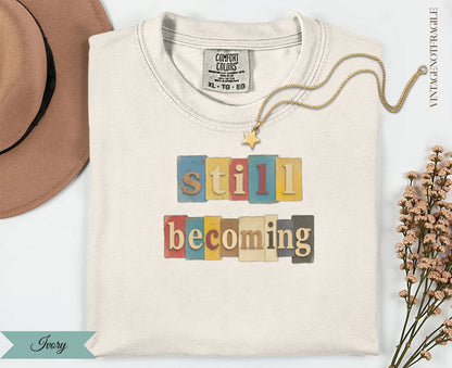 Cream t-shirt with "still becoming" text in colorful block letters.