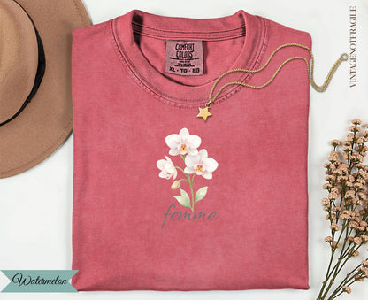 Pink t-shirt with watercolor flowers and "femme" text, gold necklace and hat nearby.
