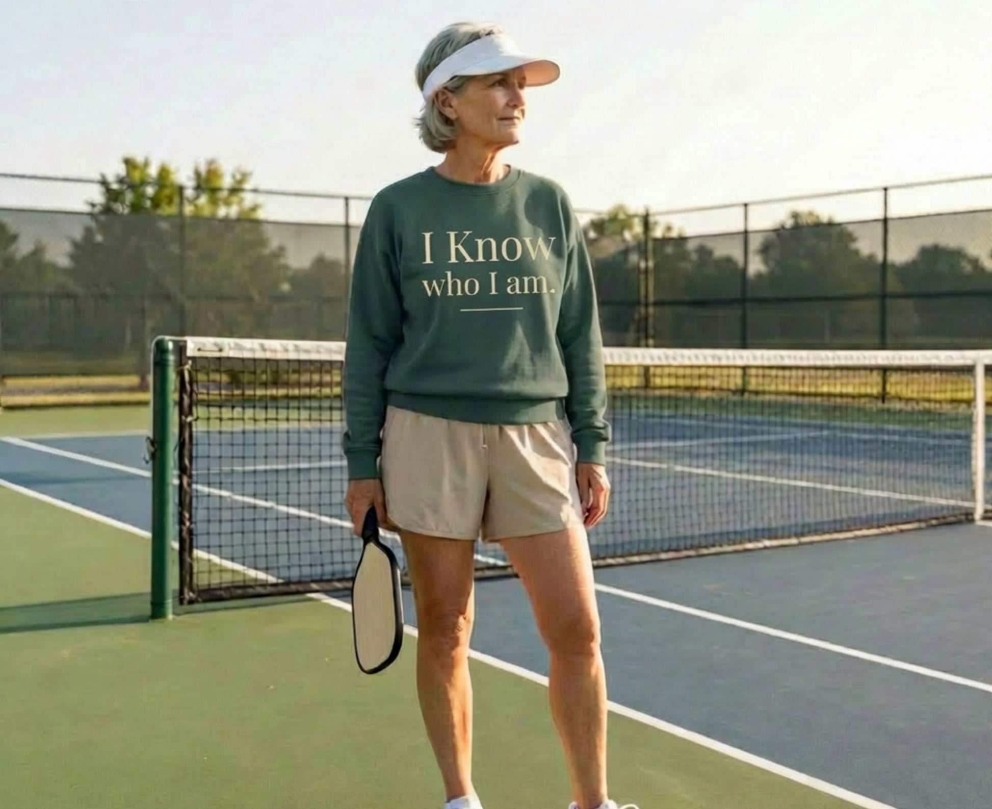 Woman in green sweatshirt with text "I Know Who I Am" holds pickleball paddle.