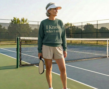 Woman in green sweatshirt with text "I Know Who I Am" holds pickleball paddle.