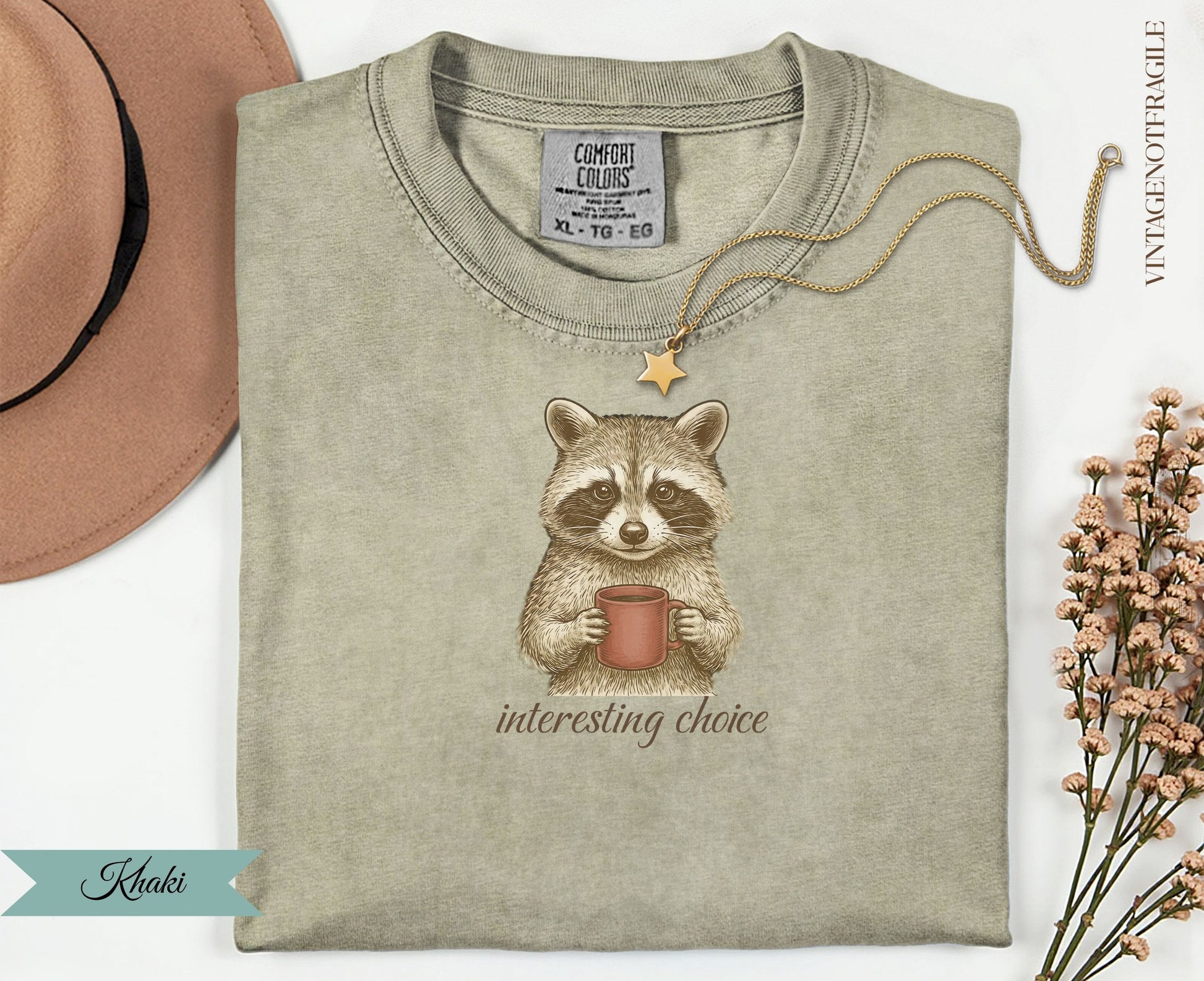 Quirky raccoon wearing a necklace holds a mug of coffee on a tan t-shirt.