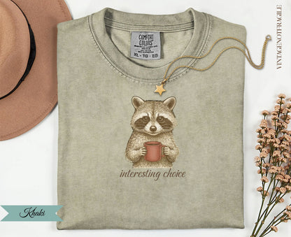 Quirky raccoon wearing a necklace holds a mug of coffee on a tan t-shirt.
