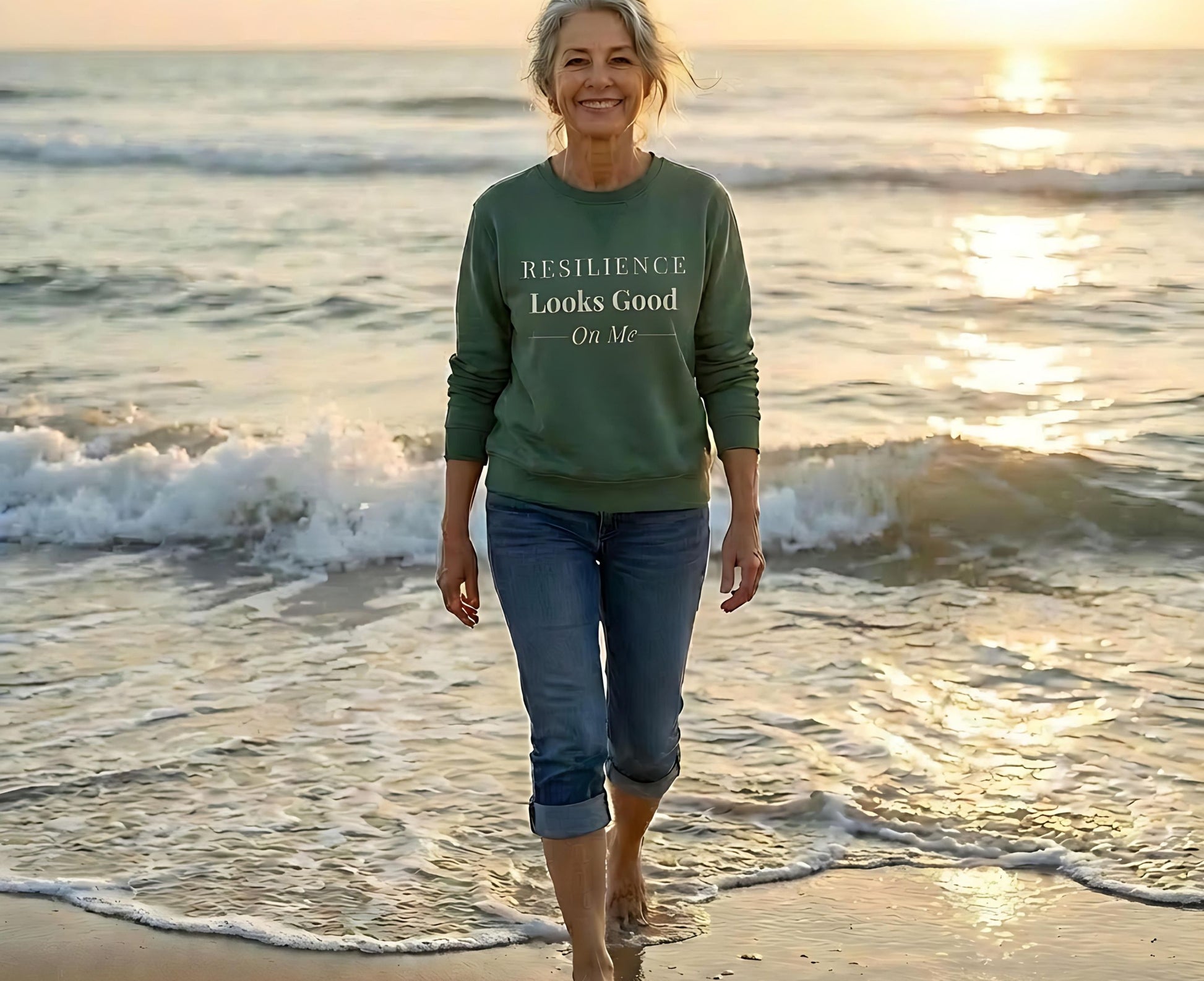 Woman walking on a beach at sunset wearing a green sweatshirt that says "Resilience Looks Good On Me."