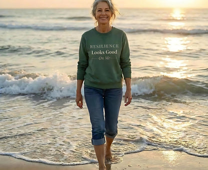 Woman walking on a beach at sunset wearing a green sweatshirt that says "Resilience Looks Good On Me."