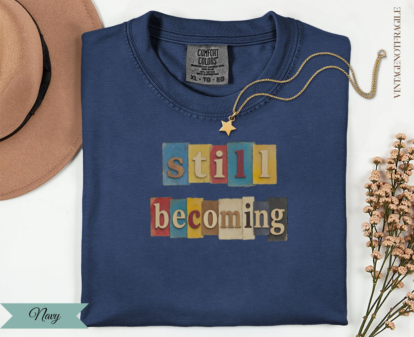Navy t-shirt with "still becoming" text in block letters on a cream background.