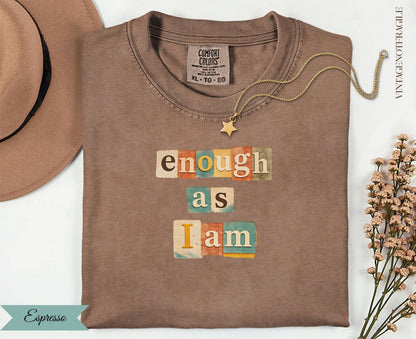 Brown t-shirt with "enough as I am" text in colorful blocks, gold necklace.