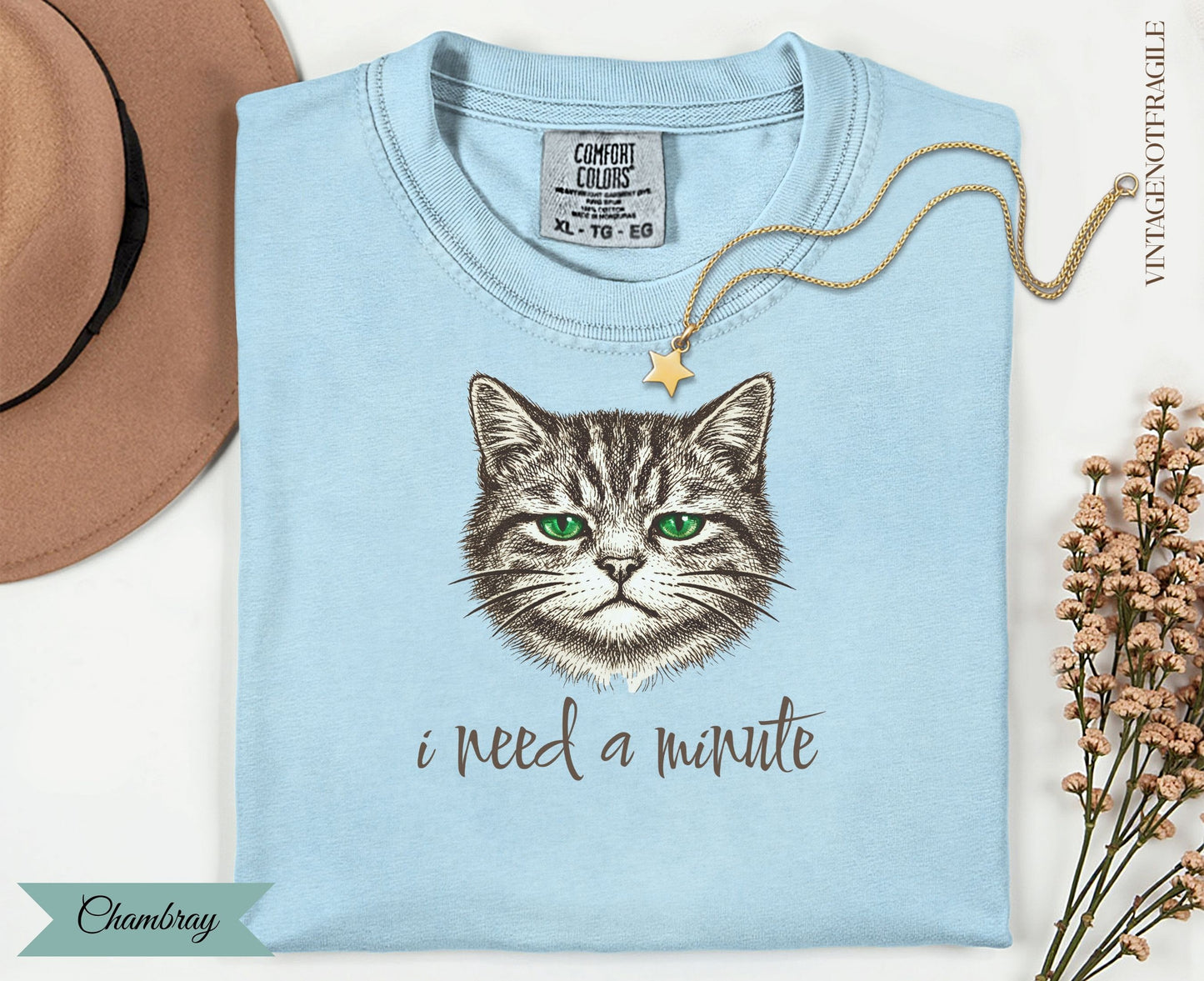 Light blue t-shirt with a graphic of a green-eyed cat and text "i need a minute."