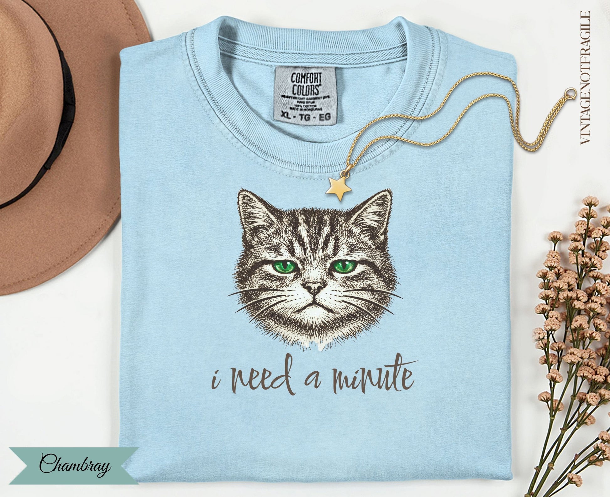Light blue t-shirt with a graphic of a green-eyed cat and text "i need a minute."
