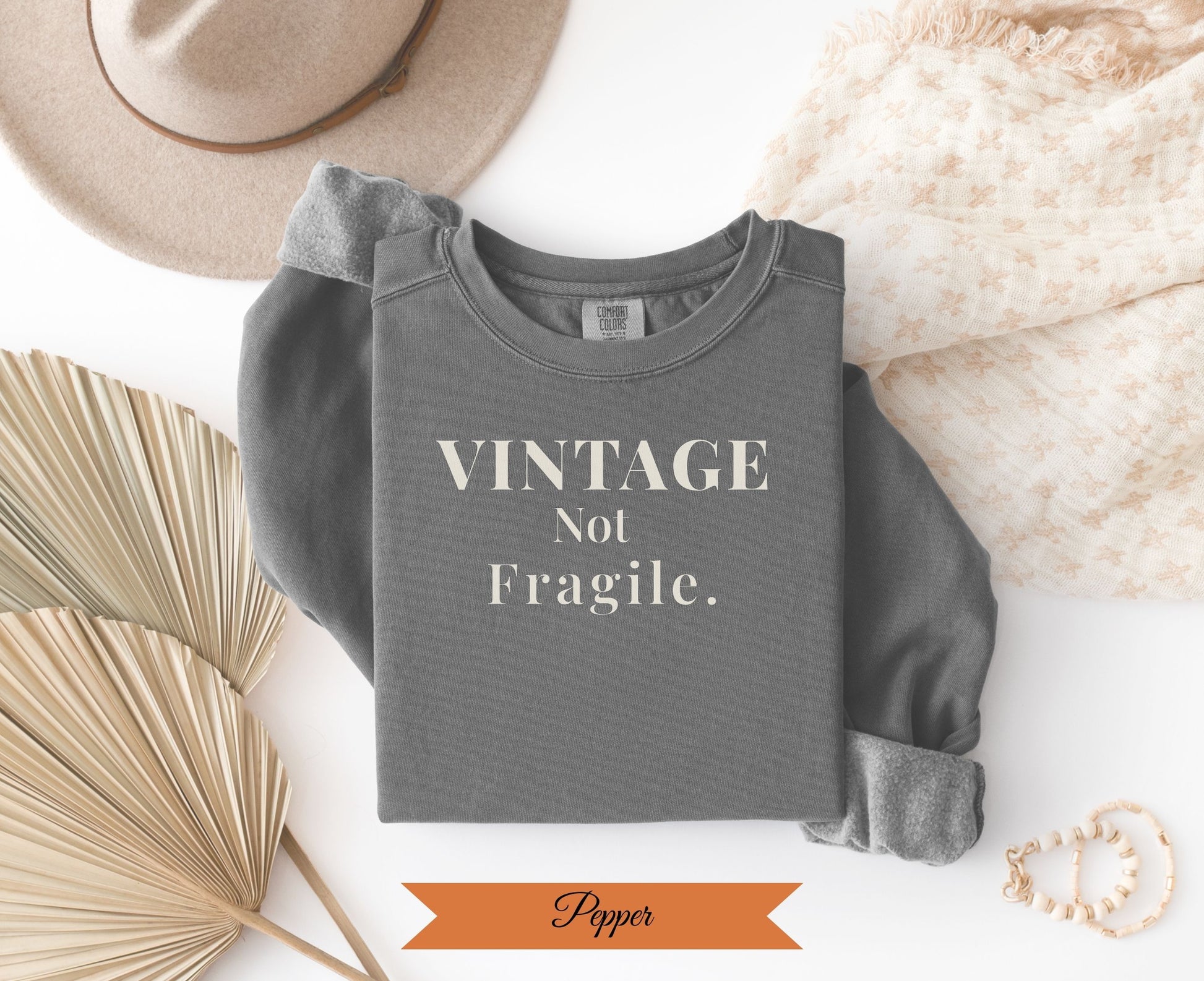 Dark grey sweatshirt with "VINTAGE Not Fragile" text, laid flat with hat and fan.