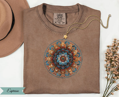 Brown t-shirt with colorful mandala design, gold necklace, and tan hat.
