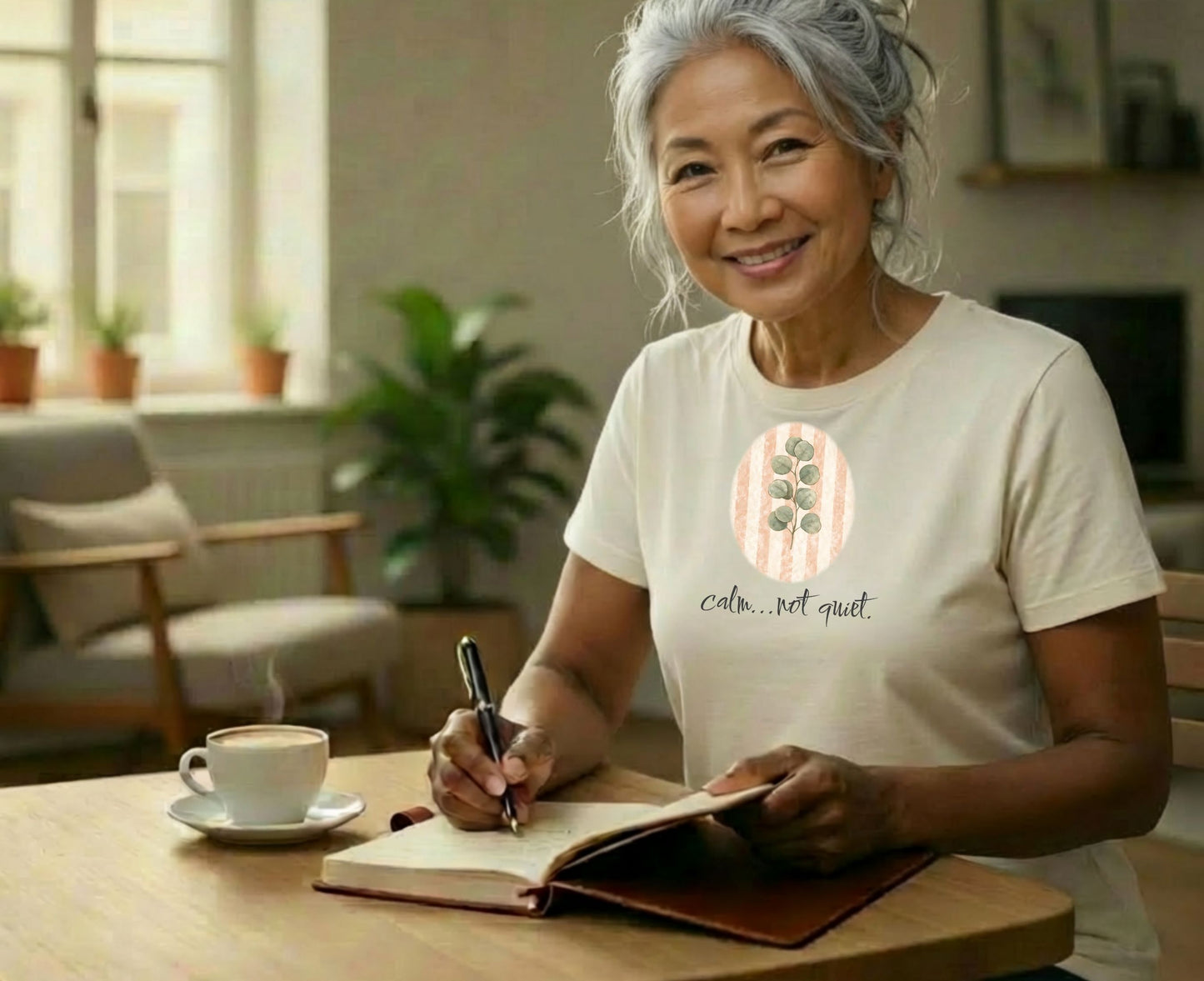 Woman with gray hair smiles while writing in a notebook with a cup of coffee.