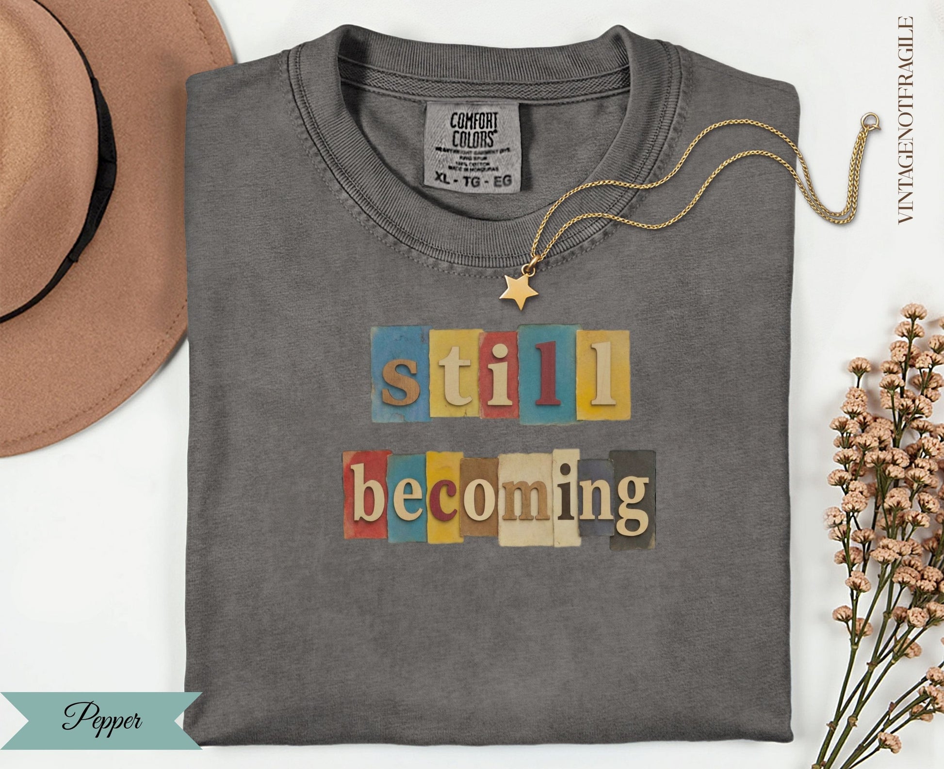 Still Becoming text on a charcoal grey t-shirt with a tan hat and gold necklace.