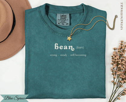 Teal t-shirt with "bean (ban)" and "strong steady. still becoming" text.