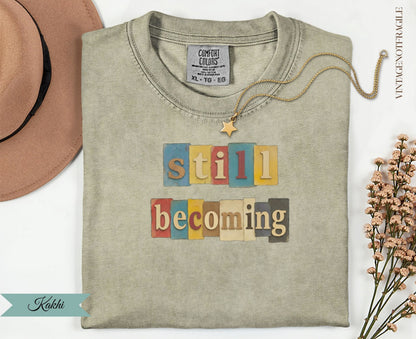 T-shirt with "still becoming" in colorful block letters.