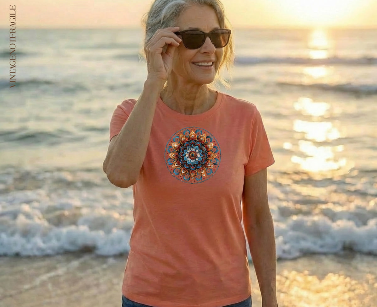 Woman in coral t-shirt with circular mandala design on beach at sunset.