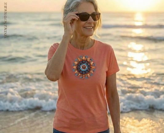 Woman in coral t-shirt with circular mandala design on beach at sunset.