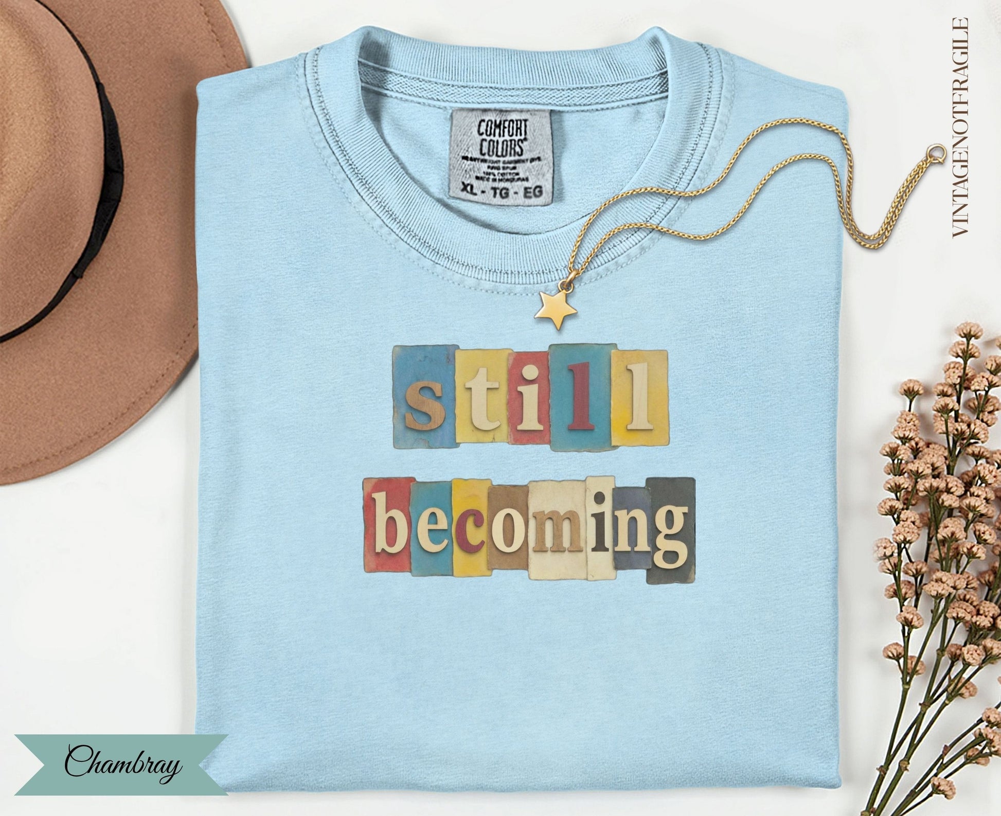 Light blue t-shirt with "still becoming" text in colorful wooden letter blocks.