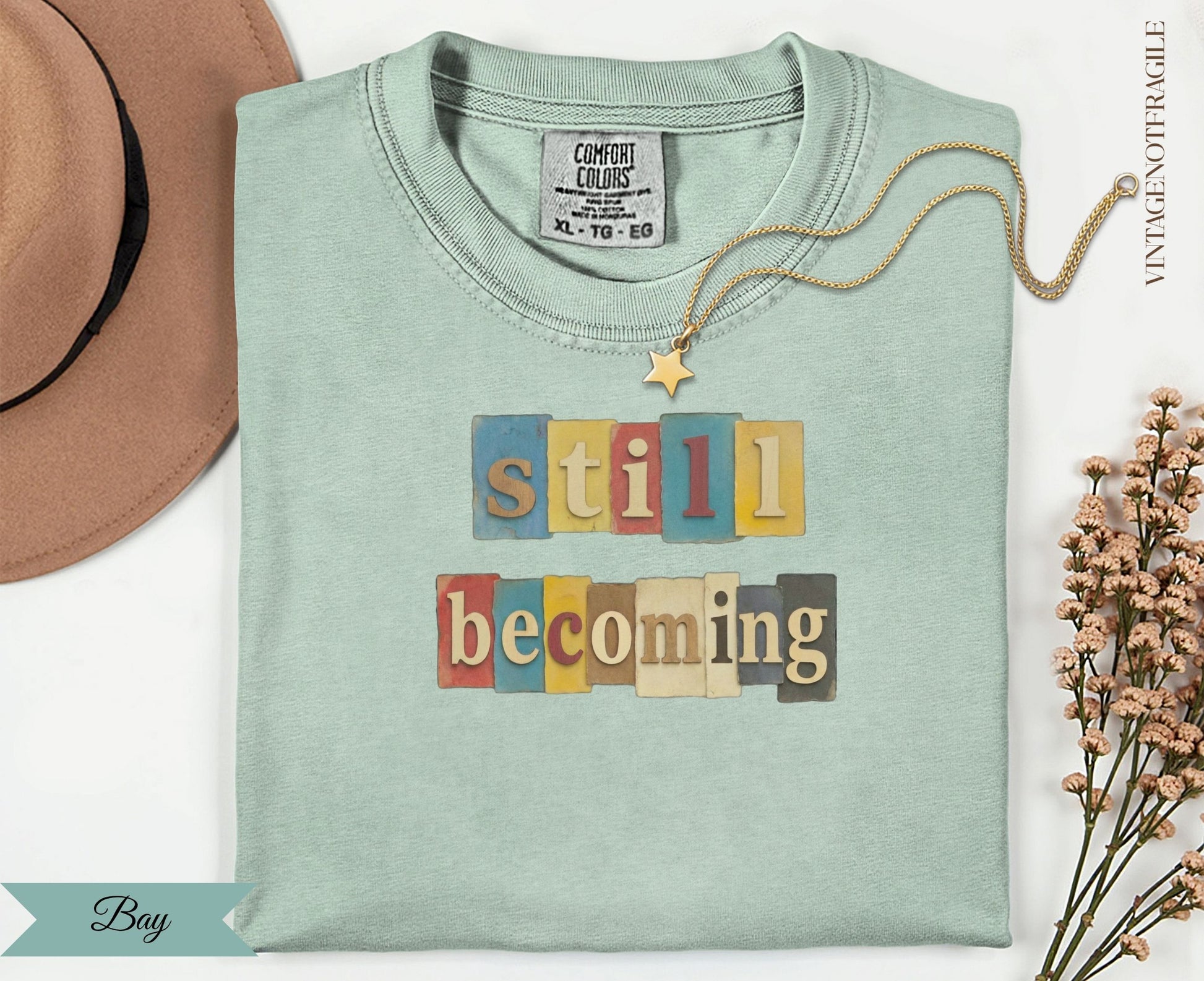 Light green t-shirt with "still becoming" text in colorful block letters.